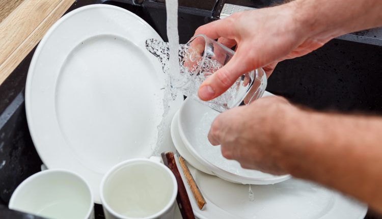 Washing dishes – man hands in gloves rinsing glass under running water in the sink