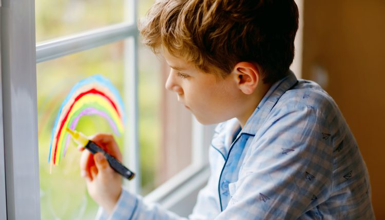 Lovely school kid boy in pajamas painting rainbow with colorful window color during pandemic coronavirus quarantine. Children painting rainbows around the world with the words Let’s all be well.