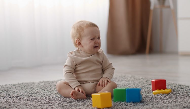 Cute little upset toddler sitting alone on floor with colorful plastic cubes and crying, looking aside for mommy