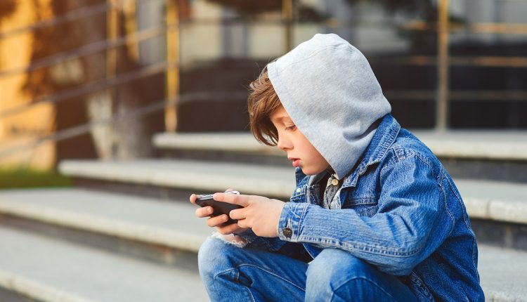 Boy use phone and plays games. Kid in the hood sitting on the stairs. Kids addicted online games and cartoons. Schoolboy plays on smartphone after school.